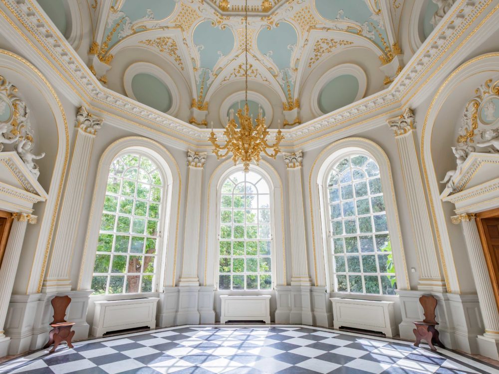 Ornate white-and-gold room with a sculpted ceiling, chandelier, tall arched windows, and black-and-white checkered floor