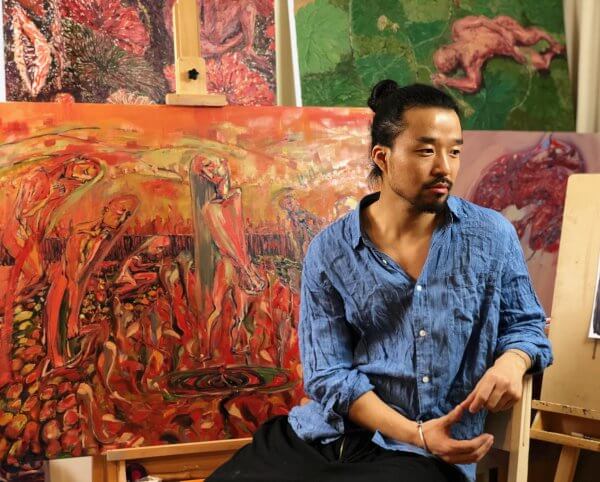 Artist Mingzhang Sun sits in a chair surrounded by several of his paintings.