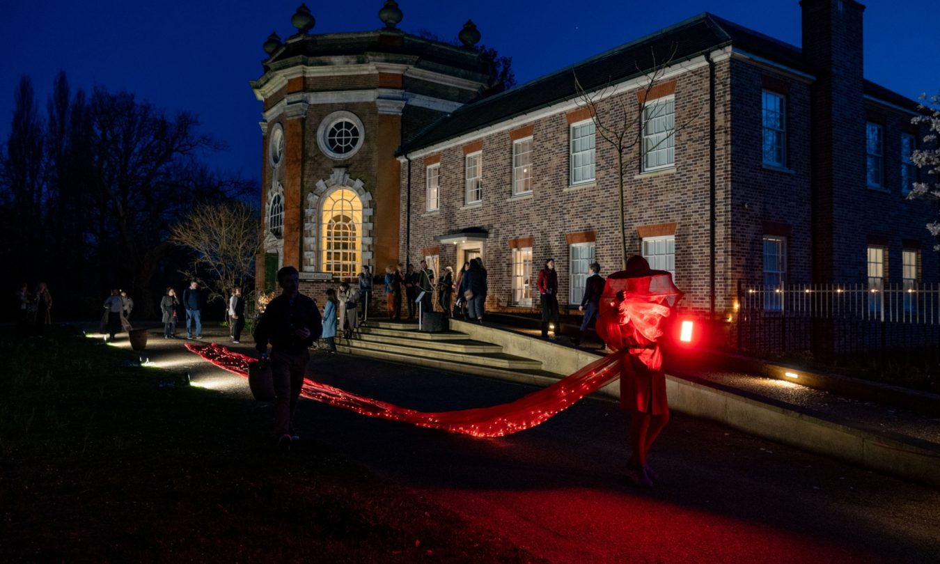 People gather outside a brick gallery at night as performers carry a glowing red fabric installation