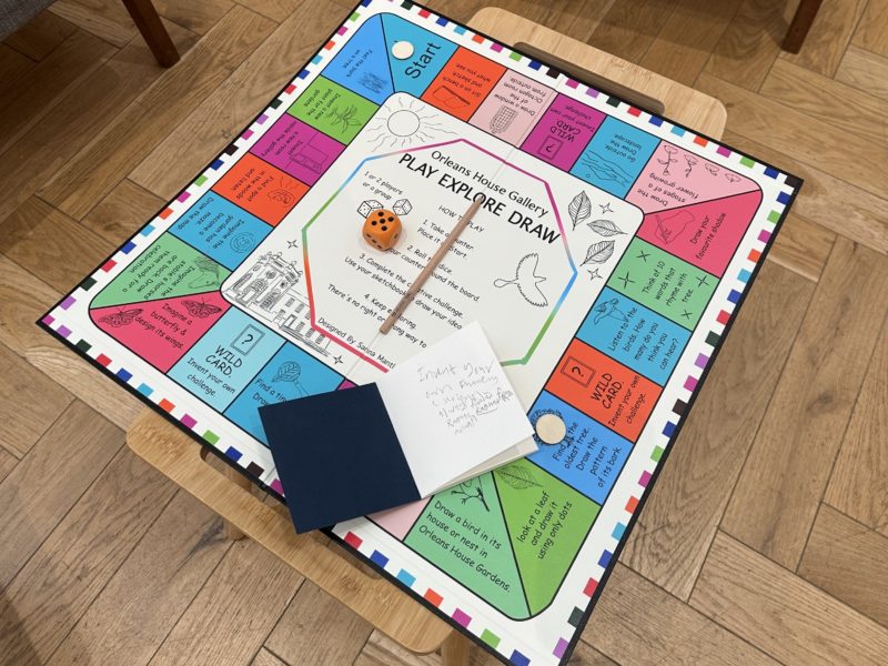 A colorful Orleans House Gallery “Play Explore Draw” board game with a die, pencil, and sketchbook on a table