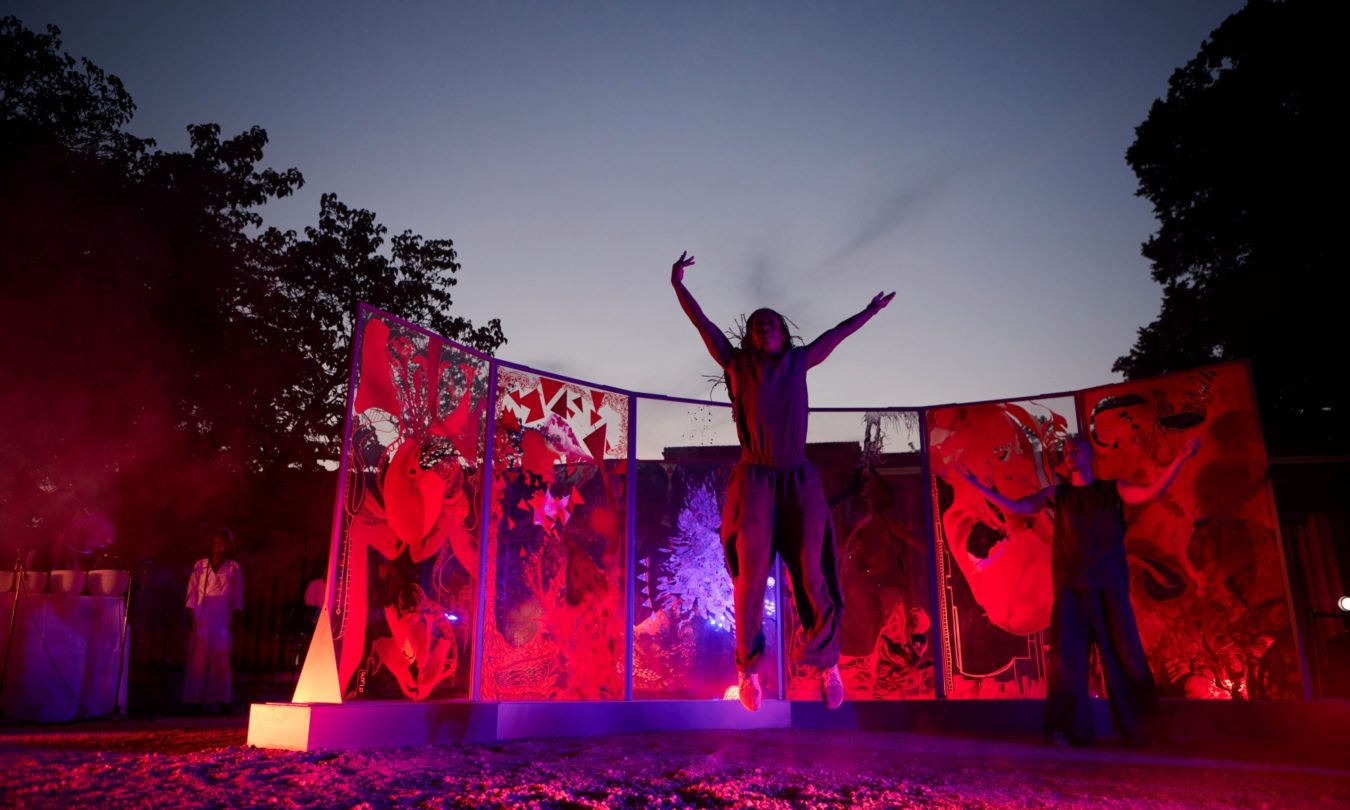 Two performers pose on an outdoor stage before colorful illustrated panels, illuminated by red and purple lights